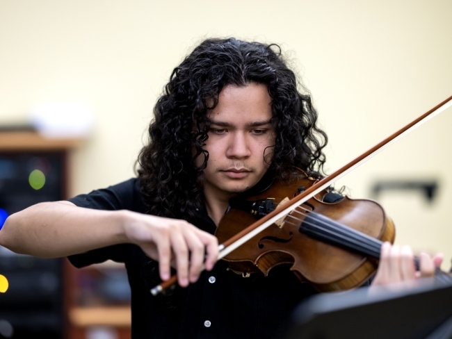A college age man with long, curly dark hair plays the violin