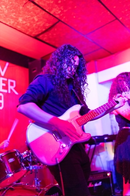 A college age man with long curly hair plays guitar onstage in vibrant pink lighting