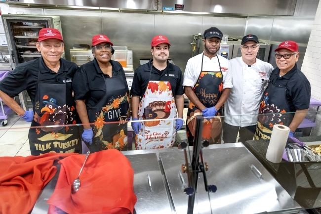 Six employees of Dining Services wear branded Davidson College hats and festive thanksgiving aprons, standing behind the serving counter