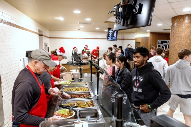 Faculty and staff wear funny hats and aprons while serving food in Vail Commons