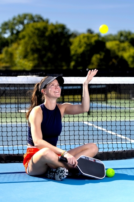 a young woman sits on a pickleball court while throwing a ball in the air and smiling