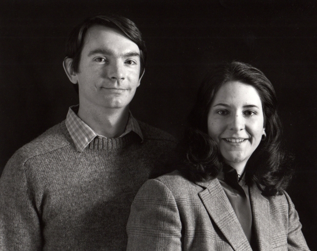 A black and white studio portrait of a man in a crewneck sweater and collared shirt standing next to a smiling woman in a patterned blazer.