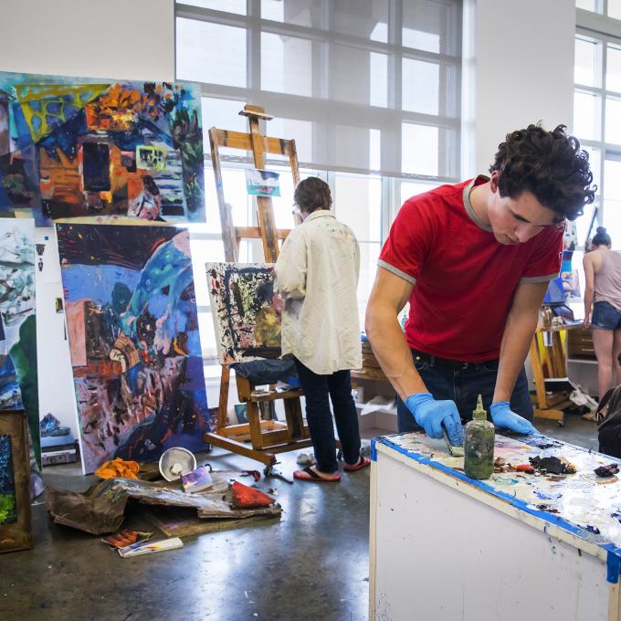 Student mixes paint in foreground while other students paint canvases in background