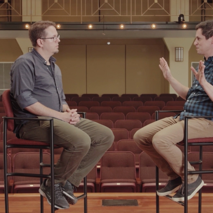 Two men facing each other in chairs onstage