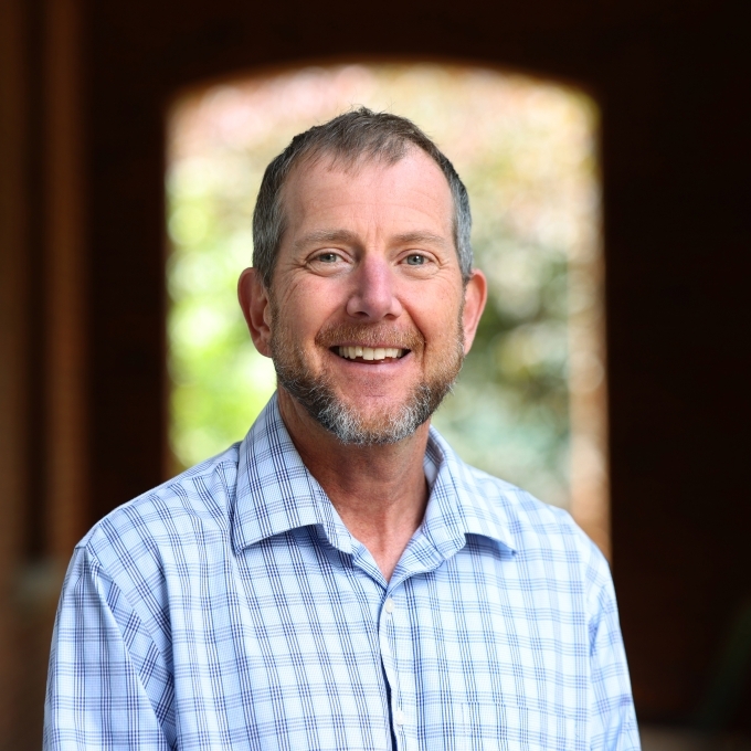 A white male wearing a blue and white gingham shirt smiles and stands with an arch behind him