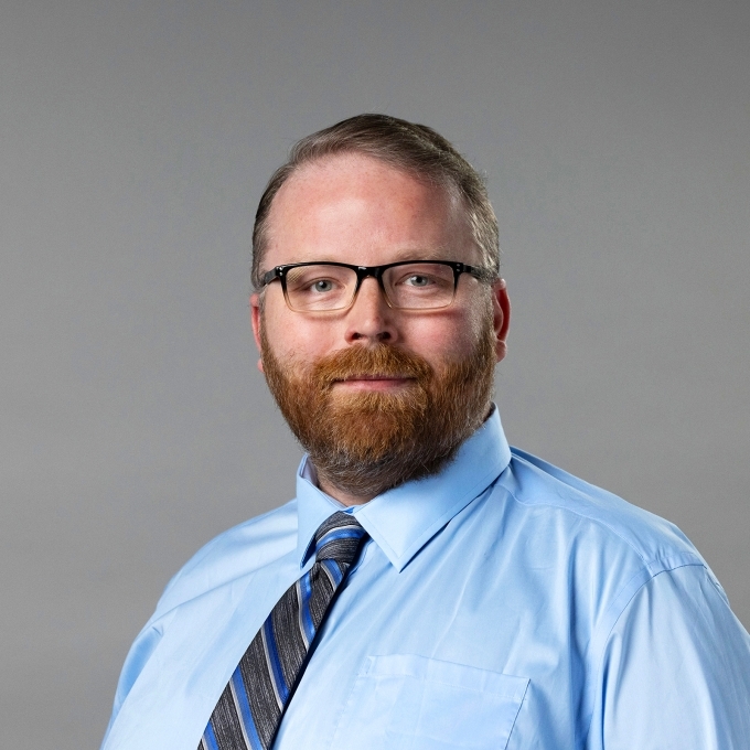 a middle aged white man with red hair and facial hair wearing glasses, a blue shirt and a tie