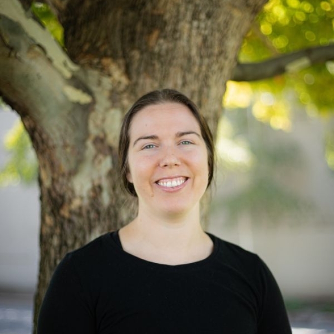 a young white woman with dark hair wearing a black top standing outside in front of a tree and smiling
