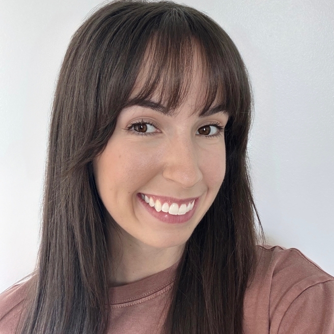 a woman with long straight dark hair smiling wearing a pink top on a grey background