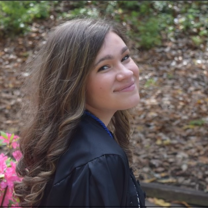 a woman with curly brown hair wearing a black jacket smiling in front of a bed of mulch and flowers