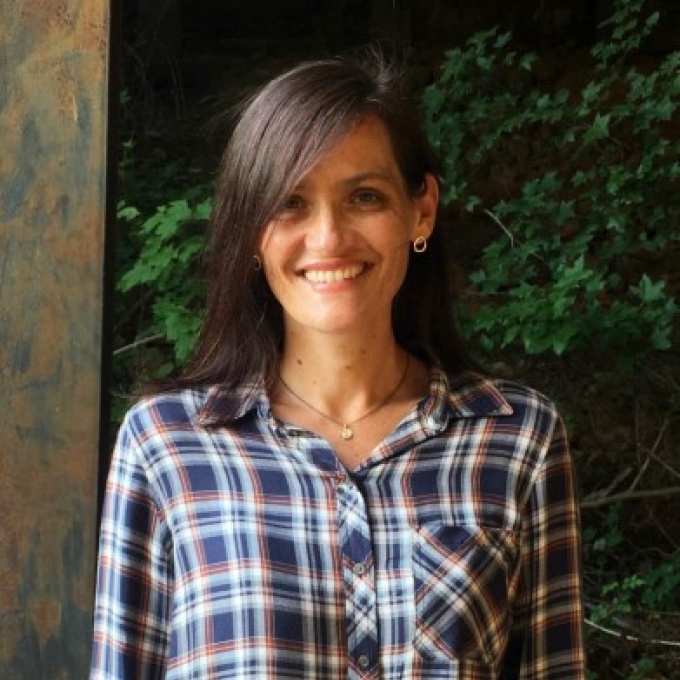 a woman with dark wavy hair wearing a flannel top in front of a green plant