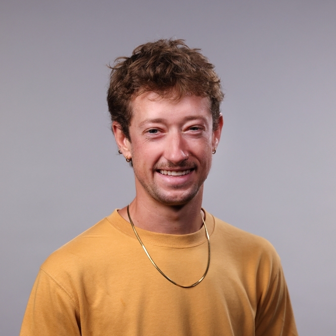 a professor wearing a mustard yellow sweater and gold chain with curly dirty blonde hair