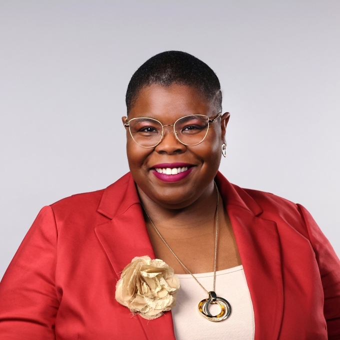 a Black woman wearing glasses, a red blazer and gold jewelry on a grey background