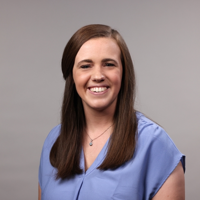 a woman with short brown hair wearing a purple blouse on a grey background