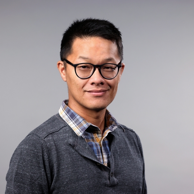 a man with short black shair wearing glasses, a flannel and half zip sweater on a grey background