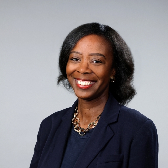 a woman with curly dark hair wearing a blazer and smiling on a grey background