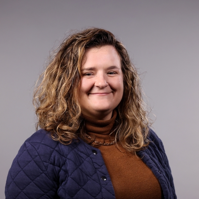 a woman with curly dirty blonde hair wearing a burnt orange turtleneck and quilted jacket on a grey background