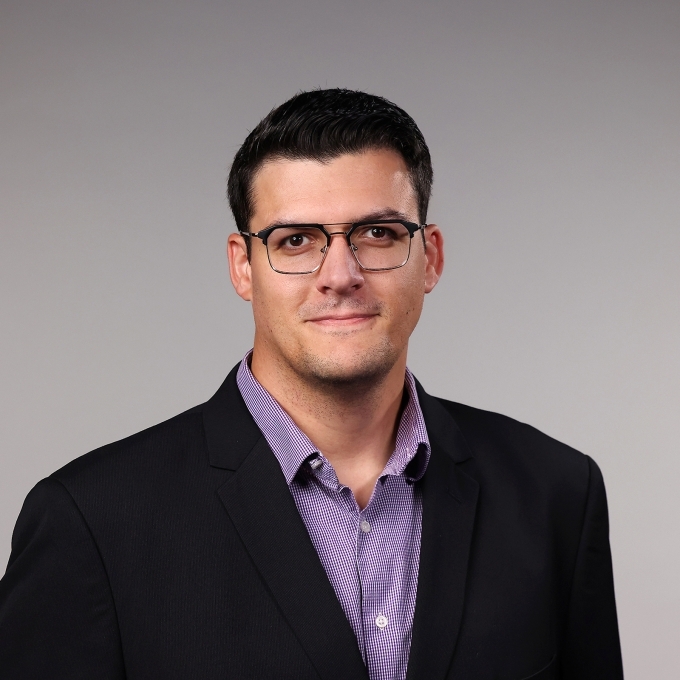 a man with short dark hair wearing glasses, a purple collared shirt and a black blazer on a grey background