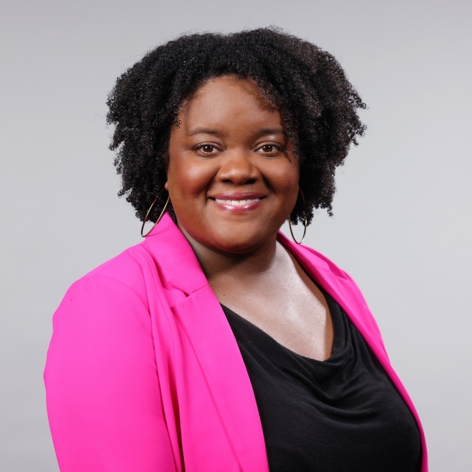 a woman with curly dark hair wearing a black top and pink blazer over a grey background