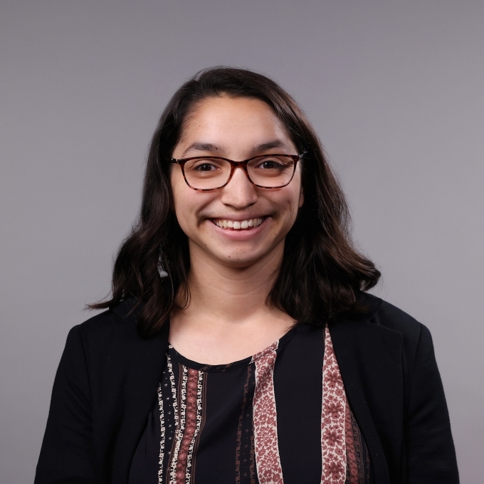 a woman with dark hair wearing red glasses and a dark blouse on a grey background