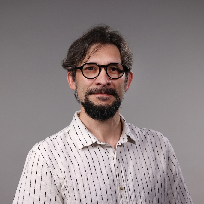 a man with dark hair and facial hair wearing glasses and a white polo on a grey background