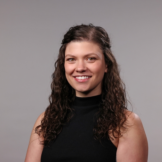 a woman with curly brown hair half-pinned back wearing a black turtleneck and smiling in front of a grey background