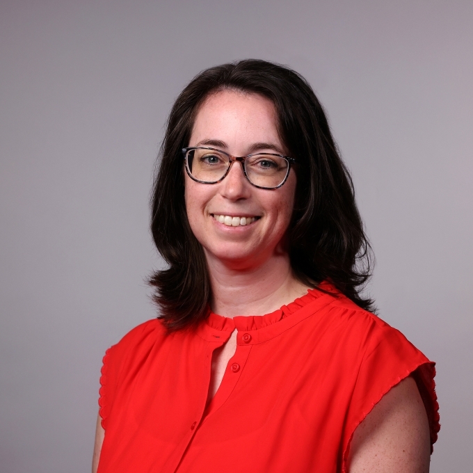 a woman with curly brown hair wearing glasses and a red top on a grey background