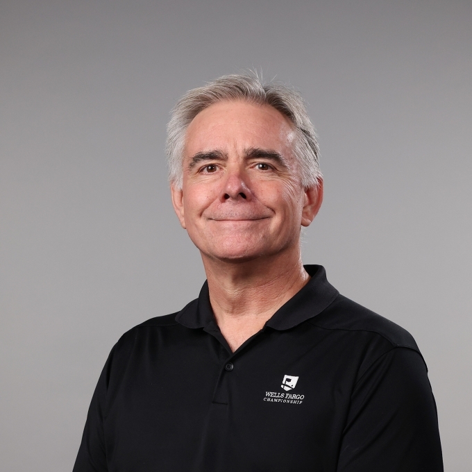 a man with short white hair wearing a collared black shirt and smiling on a grey background