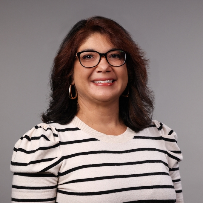 a woman with dark curly hair wearing glasses and a striped top on a grey background