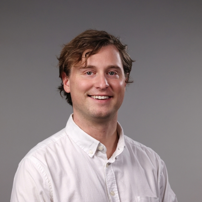a professor with short dirty blonde hair wearing a white collared shirt smiling on a grey background