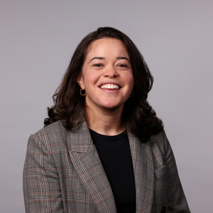 a professor with curly dark hair wearing a black top and grey blazer on a grey background