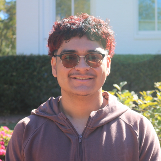 a young man with reddish brown hair wearing glasses and a sweatshirt standing outside on a sunny day