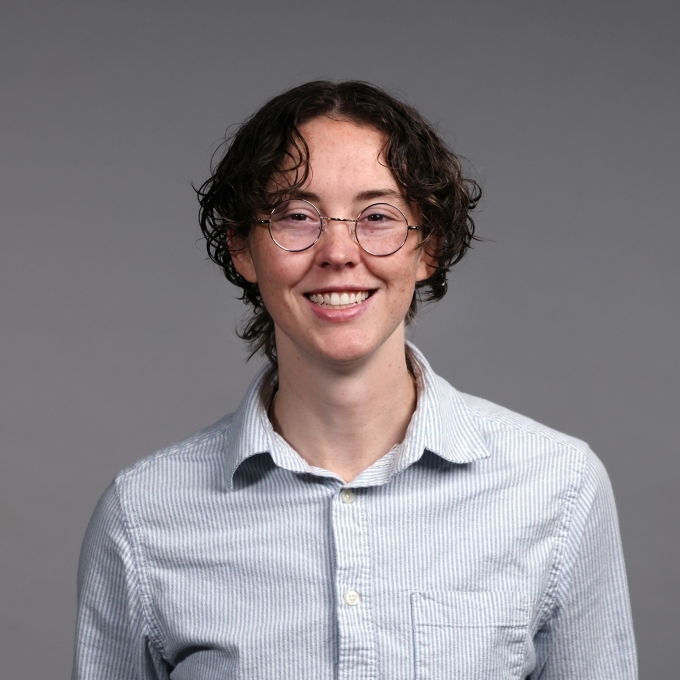 a young person wearing a collared shirt with short curly hair and glasses on a grey background