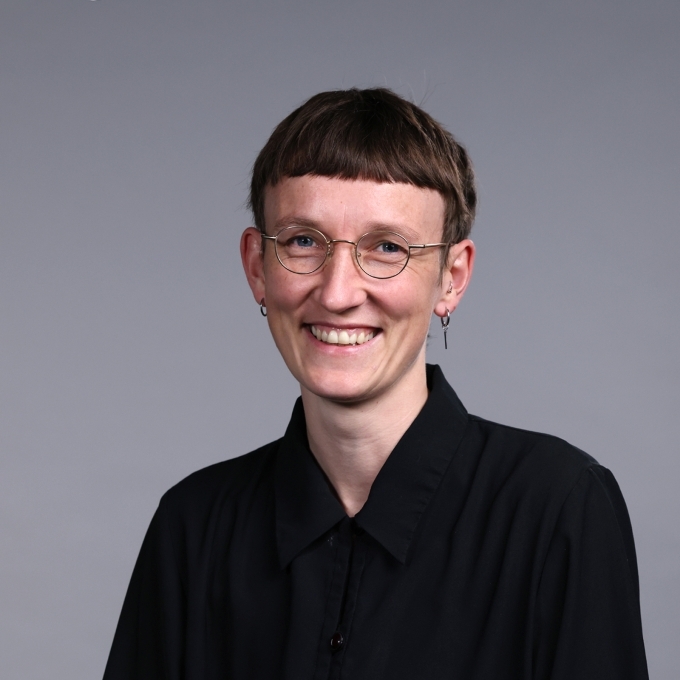 a young person with short hair wearing glasses and a black blouse on a grey background