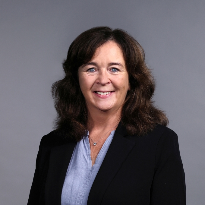 a woman with curly dark hair wearing a blue blouse and black blazer on a grey background