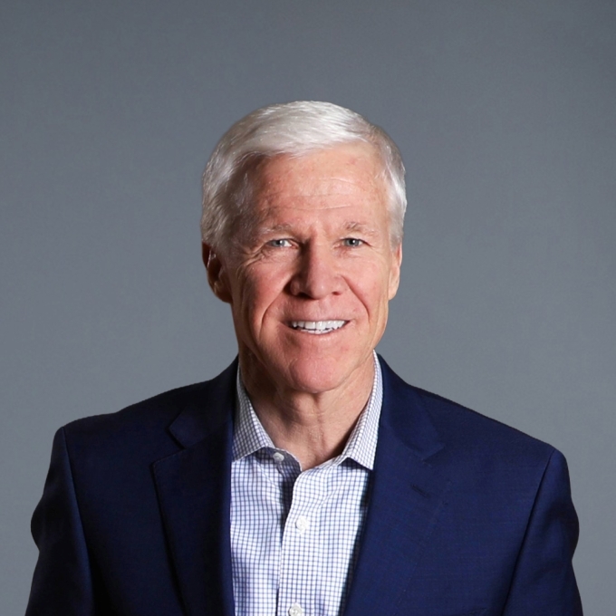 a white man with white hair wearing a collared shirt and jacket on a grey background