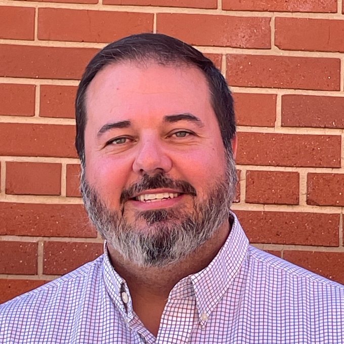 a man with facial hair wearing a collared shirt in front of a brick wall
