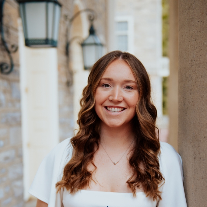 A smiling young woman with long, wavy brown hair stands outdoors wearing a white dress and holding a navy blue graduation cap with a white tassel.