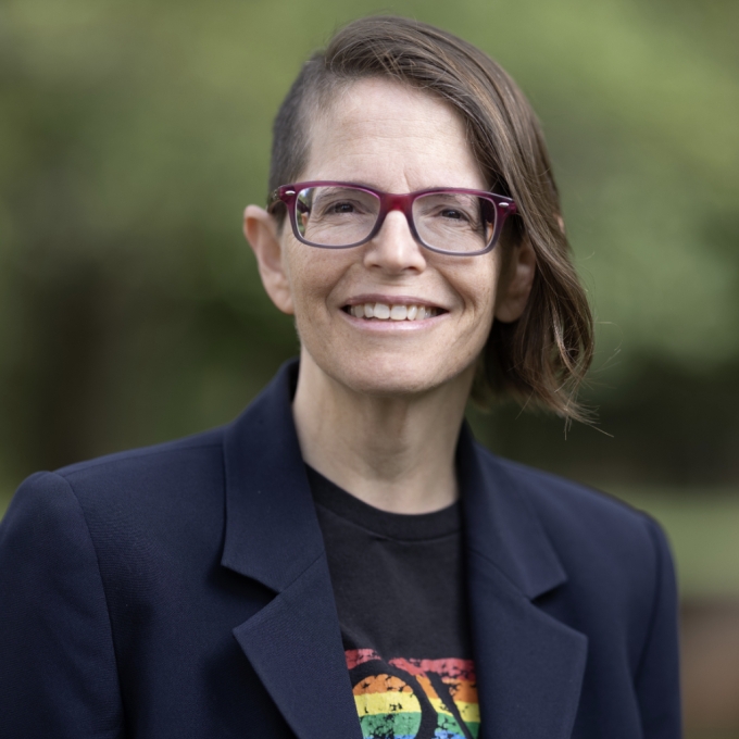 A person with brown, wavy hair in an asymmetrical style wears a blue blazer, glasses, and graphic tee