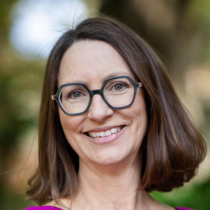 A woman with shoulder-length brown hair and glasses and a bright purple shirt