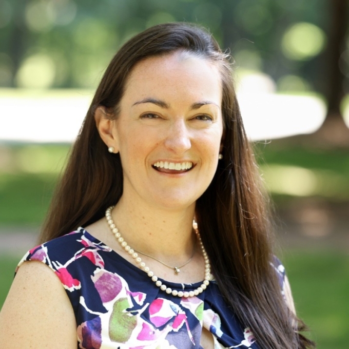A smiling woman with long brown hair, pearl earrings, and a pearl necklace, wearing a multi-colored floral sleeveless top against a blurred green outdoor background.
