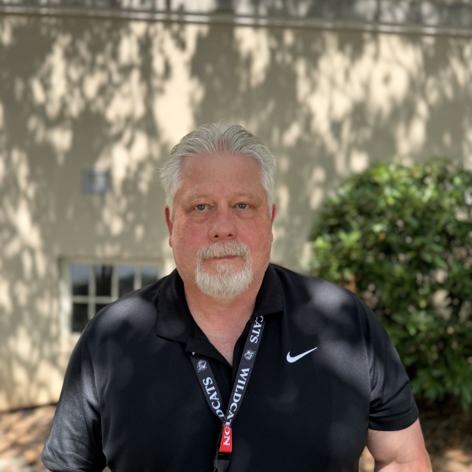 man with grey hair and a goatee wearing a black Nike polo shirt and a "Wildcats" lanyard, pictured from the waist up outdoors in front of a building.