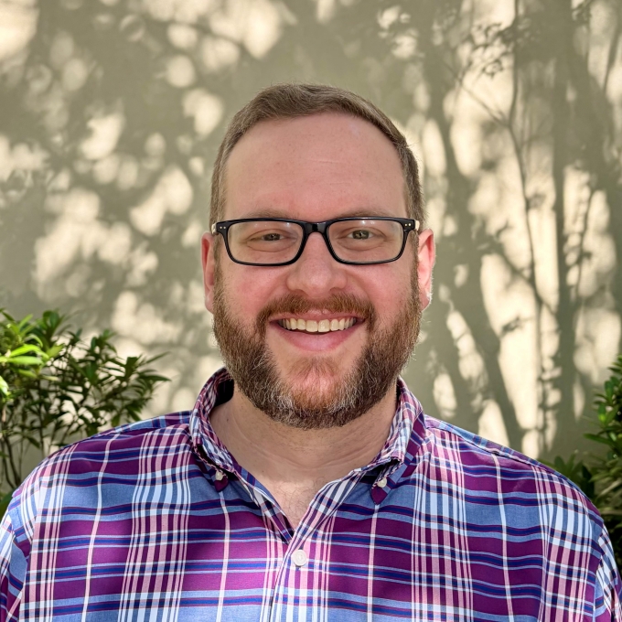 smiling man with glasses and a beard wearing a purple and blue plaid button-down shirt, pictured from the chest up against a sunlit wall with leafy shadows.
