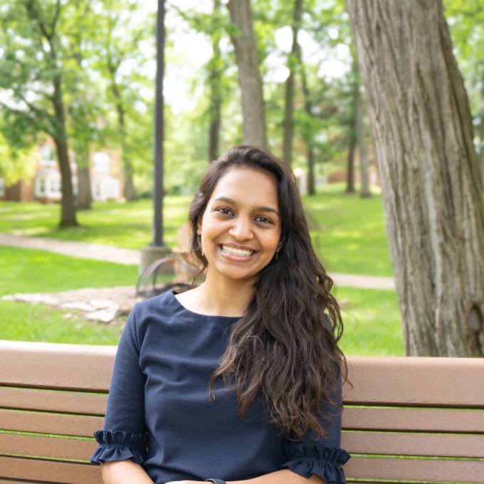 A smiling young woman with long, dark, wavy hair sits on a wooden park bench outdoors, wearing a dark blue blouse and white pants, with a green, tree-lined campus lawn blurred in the background.