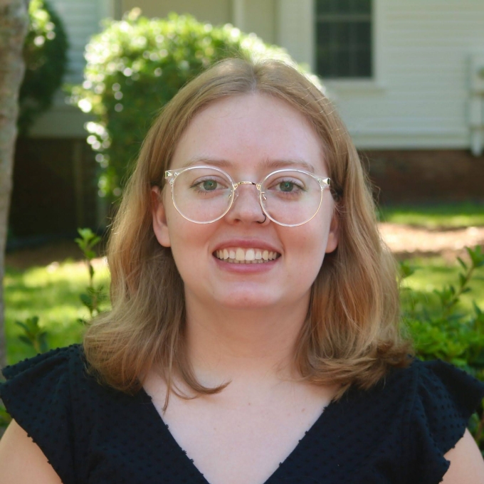 A headshot of a smiling woman with shoulder-length blonde hair, round clear-framed glasses, and a black textured top, posed outdoors against a soft-focus background of green foliage and a house.