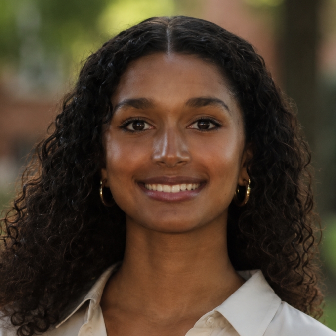 A smiling young woman with long, curly dark hair and gold hoop earrings, wearing a light-colored collared shirt against a softly blurred natural background.