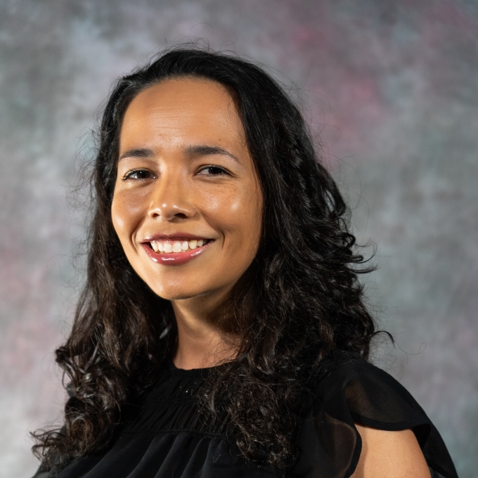 A professional headshot of a smiling woman with long, dark curly hair, wearing a black sleeveless blouse against a textured gray background.