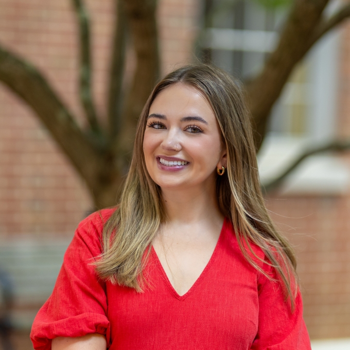 Amelia Solum smiling in a red V-neck top with a blurred brick building and tree in the background.