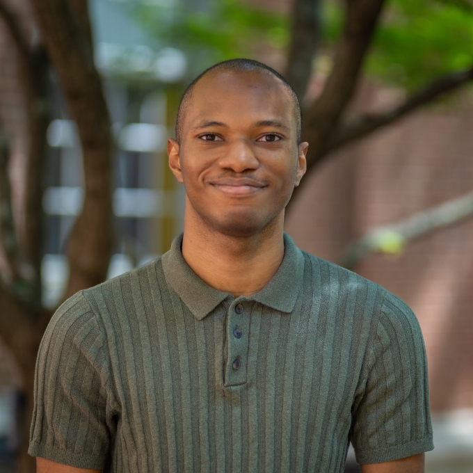 Portrait of Brillon Joseph wearing a green short-sleeved polo shirt, smiling outdoors in front of a brick building and trees.
