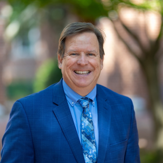 Hugh Lee smiles broadly in a professional headshot, wearing a royal blue suit jacket, light blue dress shirt, and a blue patterned tie against a softly blurred outdoor campus background.
