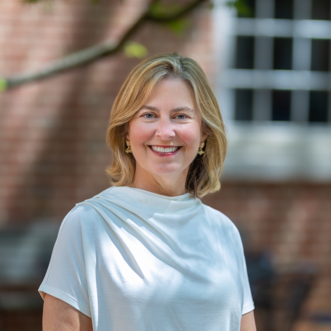 Portrait of Jane Mangan smiling outdoors while wearing a white short-sleeved blouse against a soft-focus brick building background.